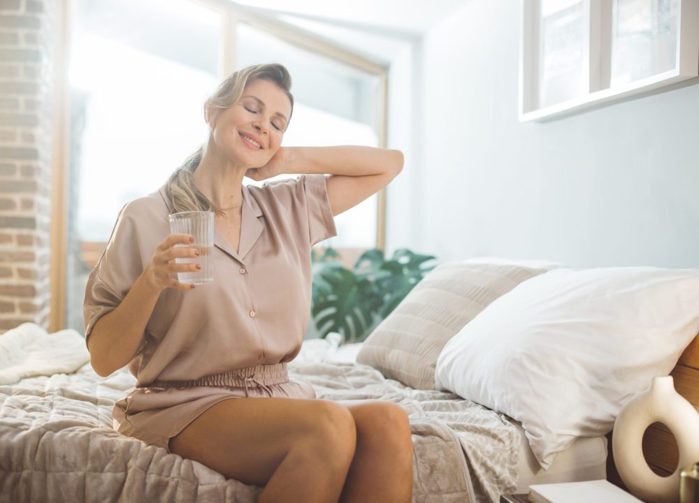 Mature woman waking up and using products for hormone replacement therapy. She is sitting on bed in pajamas and drinking water.